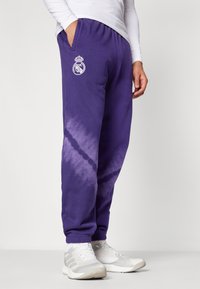 Calças de fato roxas com um padrão tie-dye, apresentando um detalhe de logotipo branco. Tecido suave, cintura elástica e bolsos laterais.