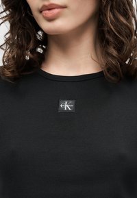 Γυναίκα με μαύρο ριπ τοπ με ένα μικρό λογότυπο Calvin Klein στη μέση κάτω από το λαιμό, με εμφανή καστανά σγουρά μαλλιά.