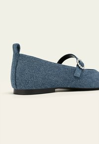 Zapatos planos de denim con punta redonda, suela negra y un cierre decorativo con un acento de strass. El color es un azul medio.