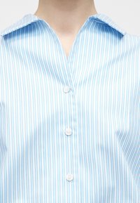 Gros plan d'une personne portant une chemise à rayures verticales bleu clair et blanc à boutons avec un col, montrant le cou et le haut de la poitrine.