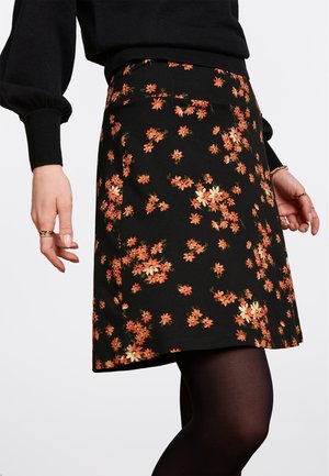 Sort nederdel med små orange og gule blomsterprint båret med sorte strømpebukser og en sort langærmet top, der viser hænder med ringe og armbånd.