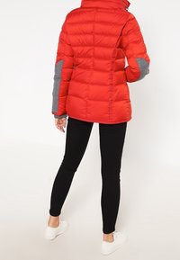 Veste matelassée rouge avec des accents gris, texture rembourrée, col montant et fermeture éclair. Portée avec des leggings noirs et des chaussures blanches.