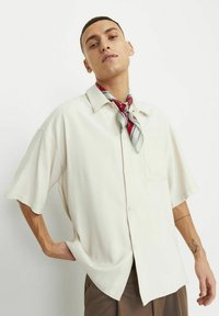 Lys beige kortærmet skjorte med knapper lavet af glat stof, med en enkelt lomme og en løs pasform, suppleret med et rødt og gråt mønster tørklæde.