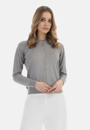 Harmaa neulecardigan, jossa on pitkät hihat, pyöreä pääntie ja etupainonappi, jonka koristeena on kultaa. Yhdistetty valkoisiin housuihin.
