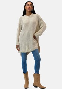 Vrouw in oversized beige gebreid trui, blauwe skinny jeans en tan suède cowboylaarzen, staand met één hand in de zak.