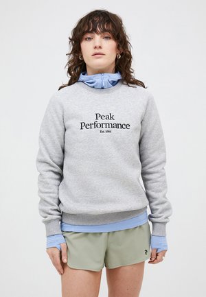 Grijze sweatshirt met "Peak Performance" in zwarte tekst, gelaagd over een blauw overhemd, gecombineerd met lichtgroene shorts. Zacht materiaal, relaxte pasvorm.