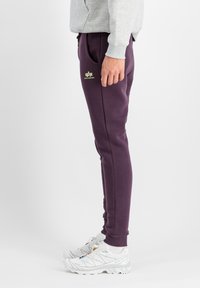 Lila Trainingshose aus weichem Stoff, mit Seitentaschen und einem kleinen gelben Logo. Kombiniert mit weißen Sportschuhen.