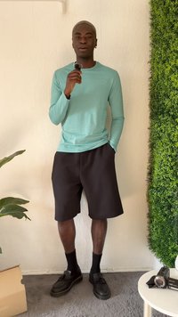 Mint green long-sleeve shirt, black shorts, black shoes, standing against a textured wall and greenery, holding a small microphone.