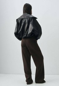 Veste oversize en cuir noir avec un large col, associée à un pantalon large marron. Texture lisse, sans motifs ni accents visibles.
