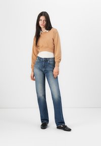 Ljusbrun kort sweatshirt med vit krage, kombinerad med blå raka jeans. Modellen bär svarta slip-on skor. Enfärgad bakgrund.