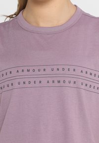 Kortärmad lavendelfärgad t-shirt i mjukt tyg, med en svart "UNDER ARMOUR" logotyp tryckt över bröstet i fet stil.