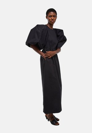 Femme portant une robe noire longue jusqu'au sol avec de grandes manches bouffantes et des sandales à talons noires à bout ouvert, debout devant un fond uni.