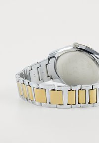 Montre argentée avec un bracelet métallique présentant des accents rectangulaires en émail jaune alternés et des maillons polis. Visage de montre rond visible.