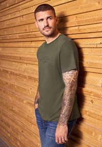 Muž s tetovanou paží, oblečený v olivově zeleném tričku a modrých džínách, stojí proti dřevěné slatové zdi a dívá se doleva.