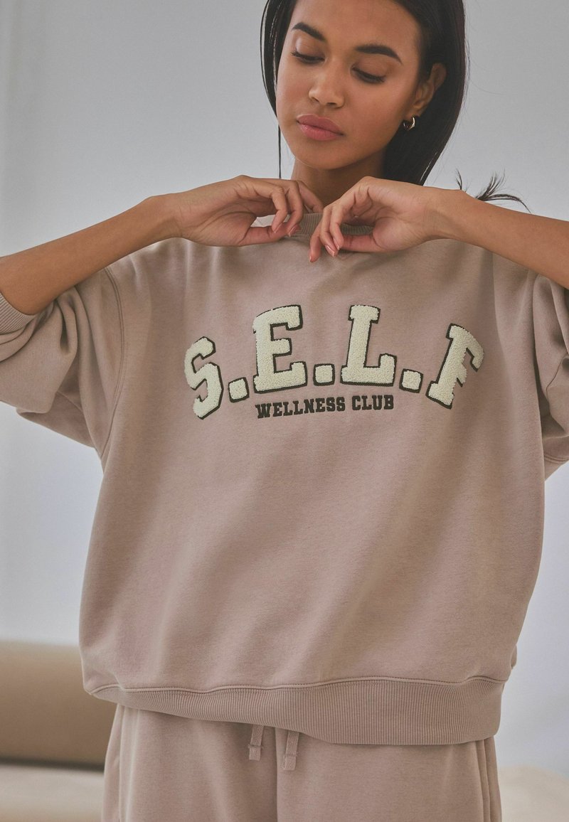 Beiger Sweatshirt mit dem Schriftzug "S.E.L.F. WELLNESS CLUB" in strukturiertem Buchstabenbild. Rundhalsausschnitt, überschnittene Schultern und gerippter Saum. Weicher Stoff.