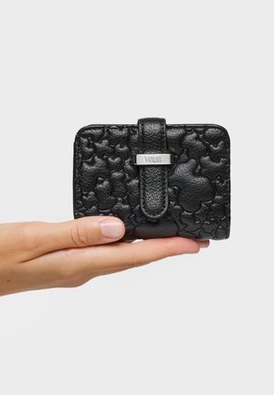 Ręka trzyma czarny skórzany portfel z tłoczonymi wizerunkami niedźwiedzi i srebrnym zapięciem z wygrawerowanym napisem "TOUS" na białym tle.