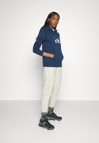 Sudadera azul marino con bolsillo frontal y logo, combinada con pantalones de chándal beige claro y calcetines estampados, zapatos deportivos negros y grises.