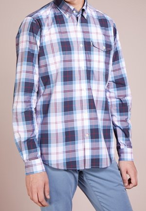 Homme portant une chemise à carreaux bleue, blanche et rouge avec des boutons, et un pantalon bleu clair, debout les bras détendus le long du corps.