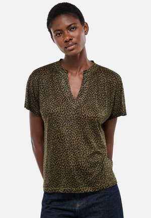 Femme noire portant un chemisier à manches courtes imprimé léopard vert olive avec un encolure en V fendue et un jean bleu foncé.