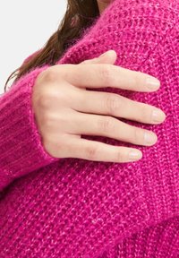 Luddig, ljusrosa tröja med en lös stickdesign. Närbild av en hand som vilar på ärmen, vilket visar dess textur och värme.