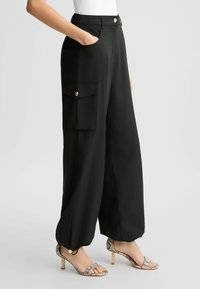 Zwarte wijde broek van gladde stof, met zijzakken en een knoopsluiting. Een opvallend detail is de cargozak met een gouden knoop.