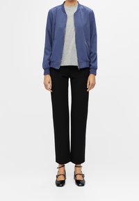 Giacca bomber blu navy con zip frontale, abbinata a una t-shirt grigia e pantaloni neri a gamba larga. Scarpe basse nere con cinturini alla caviglia completano il look.