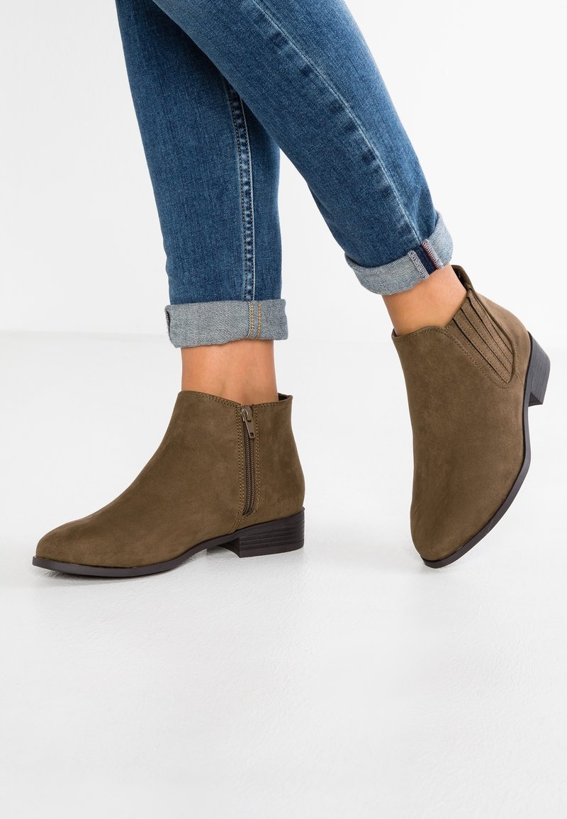 Botas de ante marrón hasta el tobillo con punta afilada y tacón bajo. Cuentan con una cremallera lateral y acentos texturizados en el eje. Combinadas con unos jeans azules remangados.