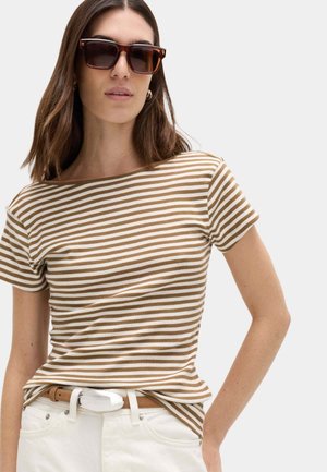 Frau trägt braun-weiß gestreiftes kurzärmeliges Hemd, weiße Hose mit beigem Gürtel und dunkle rechteckige Sonnenbrille, mit offenem Haar und goldenen Creolen.