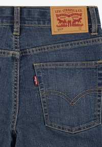Tmavomodré riflové nohavice s hnedou koženou značkovou etiketou, žltým prešívaním a červeným logom Levi's na zadnej vrecku. Klasický dizajn 511.