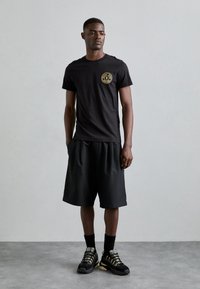 Μαύρη μπλούζα T-shirt με χρυσό κυκλικό λογότυπο, σε συνδυασμό με φαρδιά μαύρα σορτς. Μαύρα αθλητικά παπούτσια με χρυσές λεπτομέρειες και μαύρες κάλτσες. Απλή, καθημερινή εμφάνιση.