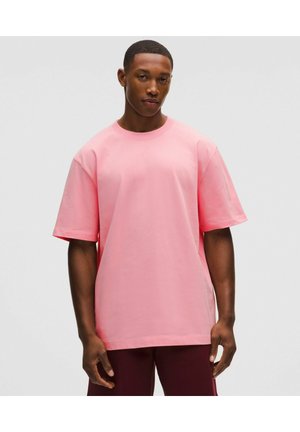 Ung mand iført en løstsiddende lyserød T-shirt med korte ærmer og mørke bukser, stående mod en ensfarvet lysgrå baggrund.