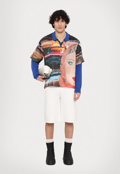 Bunt bedrucktes kurzärmeliges Hemd mit Porträt-Design, kombiniert mit weißen Shorts. Texturierte schwarze Stiefel und eine weiße Tasche in der Hand.