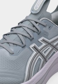 Κοντινό πλάνο ενός γκρι παπουτσιού τρεξίματος Asics με επάνω μέρος από υφασμάτινο πλεκτό με υφή, δαντέλες που διασταυρώνονται και χαρακτηριστικό λογότυπο στο πλάι πάνω σε λευκή σόλα.