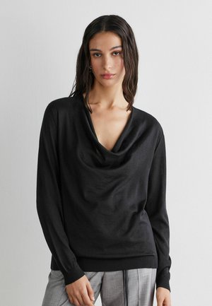 Frau mit dunklem, welligem Haar trägt ein schwarzes, drapiertes Langarmoberteil mit V-Ausschnitt und graue Hosen, steht vor einer schlichten hellgrauen Wand.