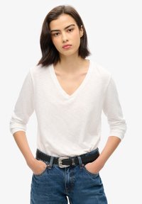 Weißes Langarm-V-Ausschnitt-Shirt aus leichtem Stoff, kombiniert mit dunkelblauen Jeans und einem schwarzen Gürtel mit einer silbernen dekorativen Schnalle.
