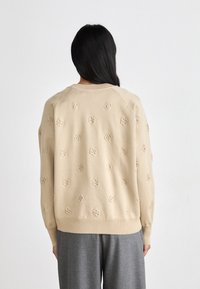 Beige pulóver kisméretű köralakú mintákkal. Lazább fazonú, bordázott mandzsettákkal és aljjal, szürke nadrággal párosítva.