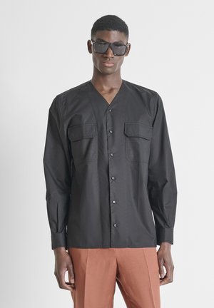 Mladi muškarac s kvadratnim crnim sunčanim naočalama, crnom košuljom dugih rukava s džepovima na prsima i svijetlosmeđim hlačama, stoji na bijeloj pozadini.