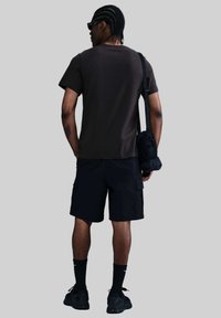 Camisa de manga corta marrón oscuro, pantalones cortos cargo negros, zapatillas deportivas negras y calcetines tobilleros negros; persona con trenzas lleva una bolsa negra.