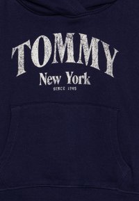 Marineblå hettegenser laget av bomullsblandet stoff, med en frontlomme og slitt hvit tekst: "TOMMY New York siden 1985."