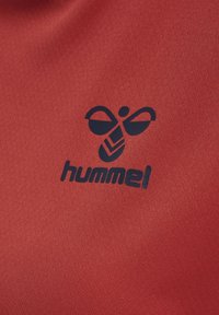 Roter Stoff mit glatter Textur, auf dem das dunkelblaue "hummel"-Logo und ein stilisiertes Insektenmuster deutlich sichtbar sind.