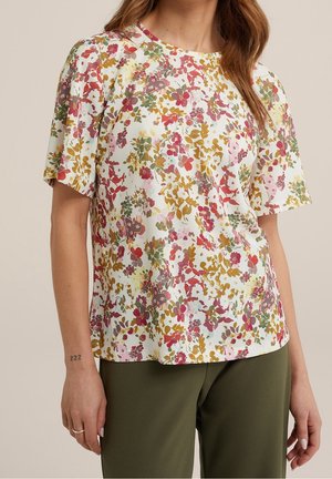 Nainen pukeutunut valkoiseen lyhythihaiseen puseroon, jossa on punainen, pinkki, keltainen ja vihreä kukkakuvio, yhdistetty oliivinvihreisiin housuihin.