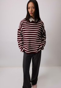 Maglione oversized a strisce bordeaux e bianche con spalle scese, abbinato a pantaloni ampi scuri e scarpe di colore chiaro.