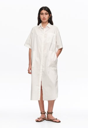 Mujer de pie con un vestido tipo camisa blanco hasta la rodilla de manga corta, con bolsillos delanteros y sandalias planas marrones con tiras.