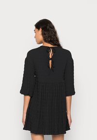 Vestido preto com pregas, apresentando um tecido texturizado, mangas três quartos e um fecho com laço nas costas. O design inclui uma cintura ajustada e uma saia evasê.