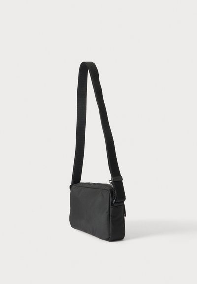 Petit sac bandoulière rectangulaire noir avec une large bandoulière réglable sur fond blanc.
