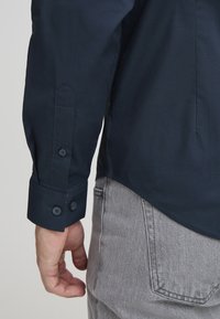 Marineblauw shirt met knoopmanchetten. Gladde stof, slim fit, schuin afgeknipte zoom, met twee donkere knopen op elke manchette. Grijze jeans op de achtergrond.