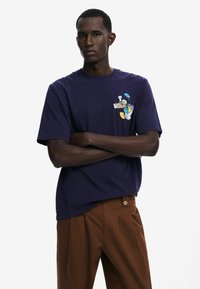 Námořnické bavlněné tričko s barevným grafickým potiskem na hrudi. Uvolněný střih s kulatým výstřihem a krátkými rukávy.