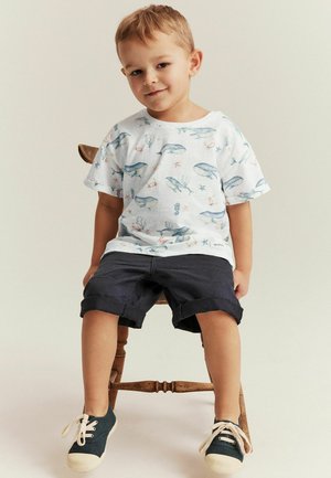 Hvid T-shirt med blå hval og havlivsmønstre, parret med mørke, rullede shorts og marineblå sneakers med beige accenter, siddende på en træstol.