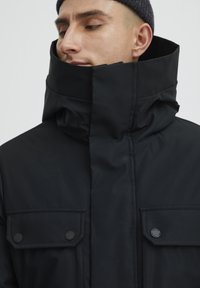Sort vinterjakke med høj krave, to lukkede brystlommer og glat tekstur. Har lynlås og minimalistisk design.