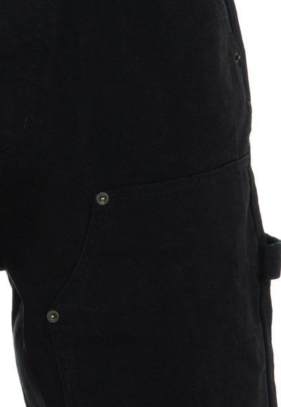 Gros plan sur un pantalon noir avec une poche latérale incurvée, deux rivets métalliques et une petite boucle en tissu.
